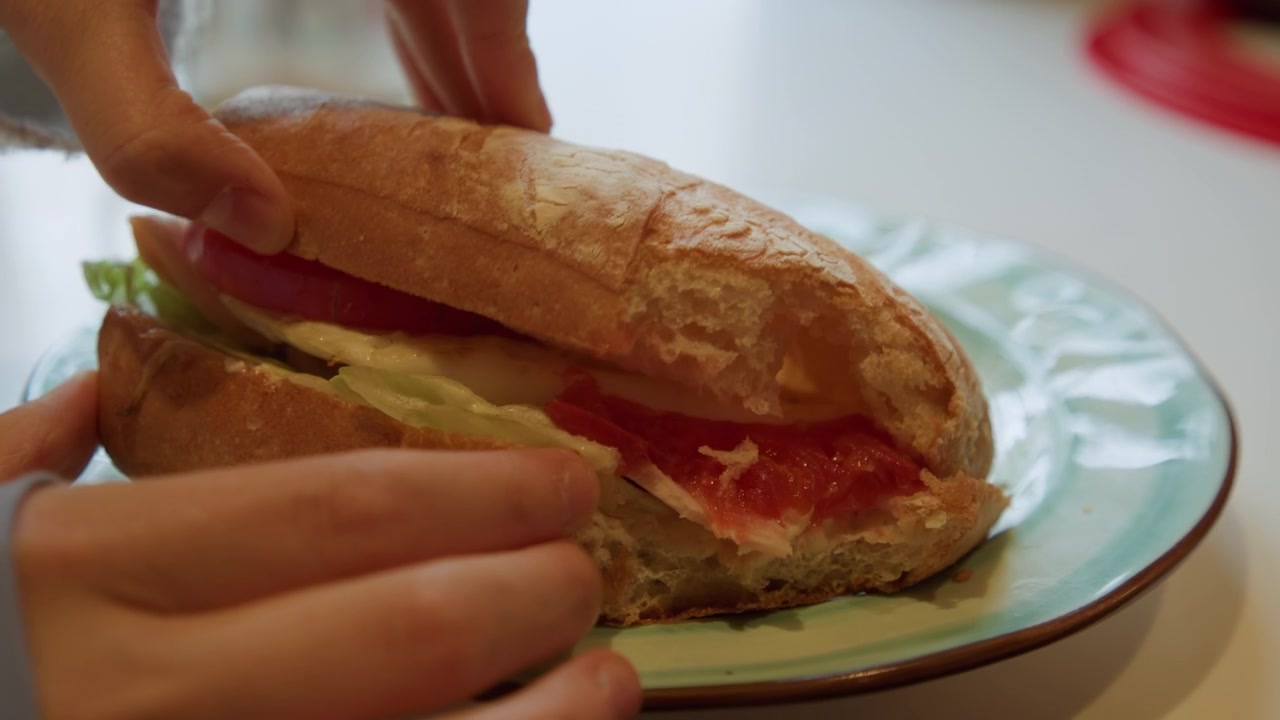 Primo piano di mani che preparano un panino con verdure fresche sul pane. Una persona maneggia gli ingredienti, posizionando pomodoro e lattuga all'interno di mezze baguette croccanti presentate su un piatto di ceramica. Soffusa...