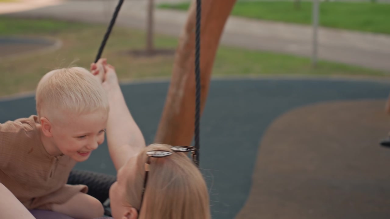 Un bambino e una donna si divertono a dondolare insieme in un parco giochi all'aperto. Il bambino si tiene all'altalena con la sua compagna, circondati da erba verde e strutture di gioco in legno