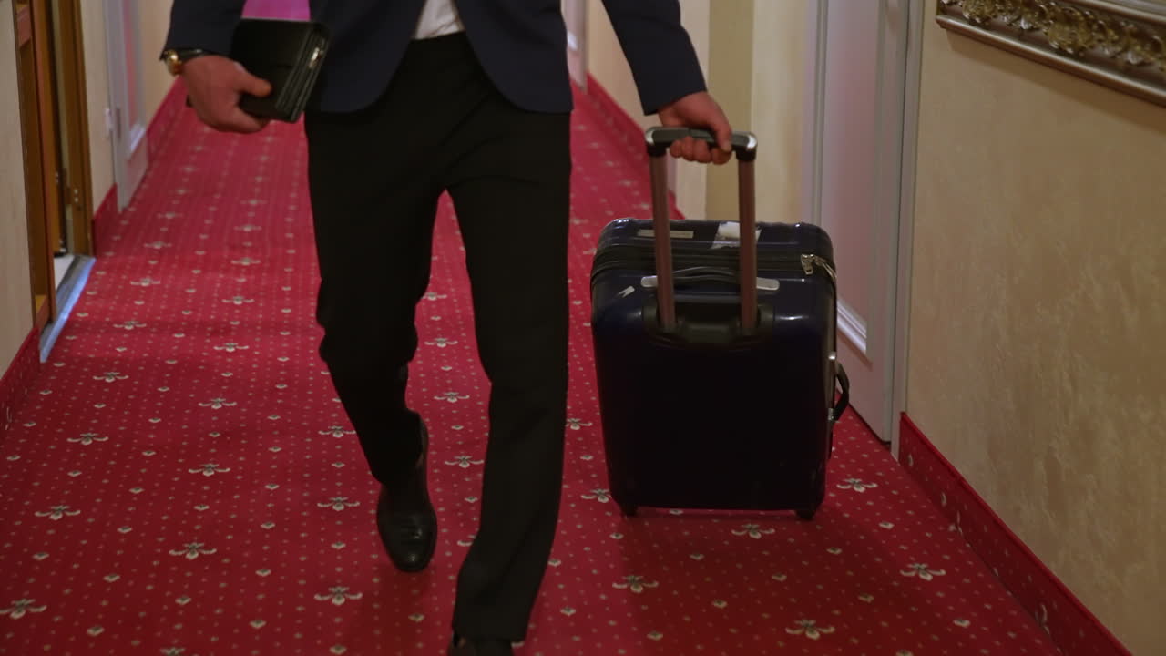 Young businessman is pulling suitcase. Travelling guy wearing elegant costume walking with his luggage in hotel hallway. Slow motion.