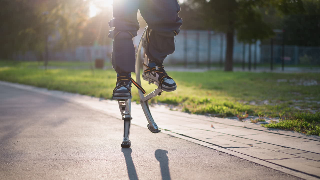 Person walking on spring stilts outdoors on paved park path, wearing dark pants and white sneakers, captured from slightly low angle. Active movement and bouncing