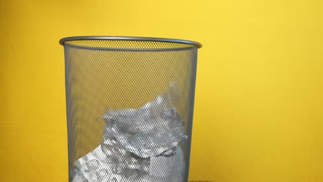 a metal trash can full of crumpled paper against a yellow background