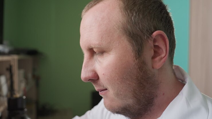 Close view of a man in a lab coat, intently focused on his work in his workshop, with a shelf in view and a green paint wall