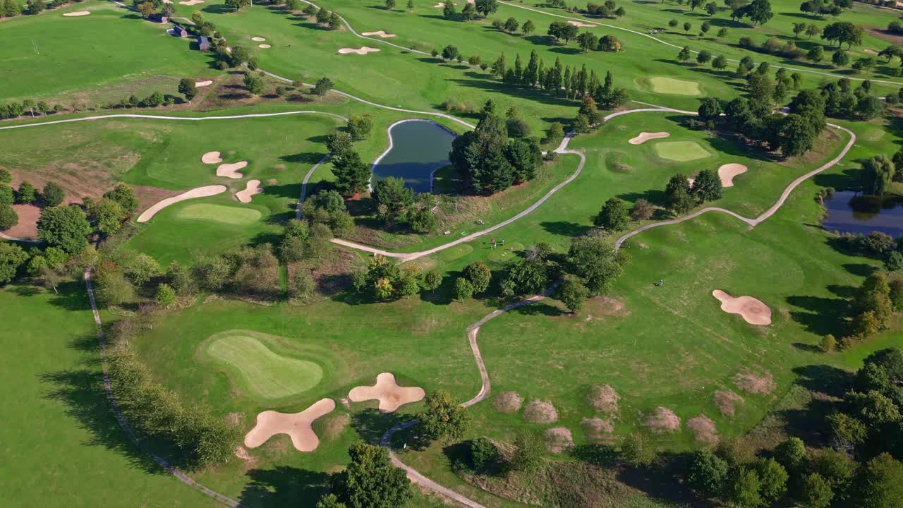 Drone flies forward above a green golf course with sand bunkers, paths, and trees under sunlight