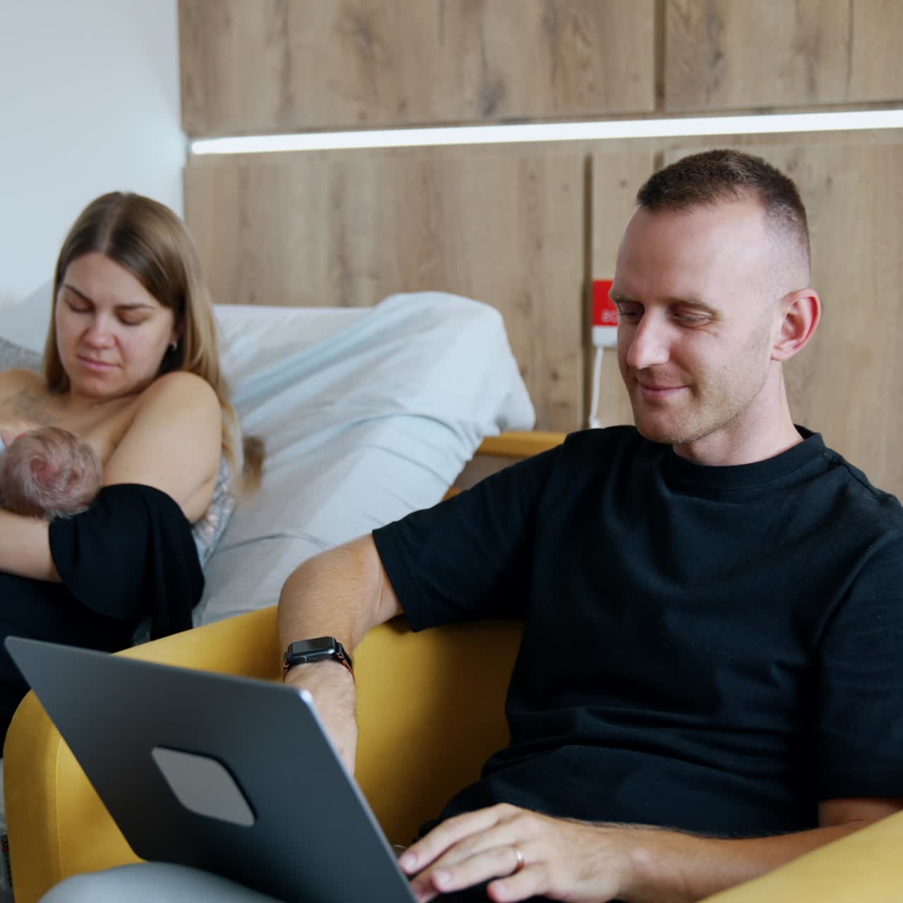 Modern family couple with a newborn in the hospital after childbirth. First time mom feeding a baby and dad listens to music on laptop