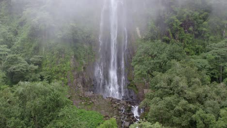 Aerial-rising-over-clouds-and-green-dense-rainforest-revealing-Las-Lajas-waterfall-streaming-into-rocky-pond,-San-Luis-Morete,-Costa-Rica