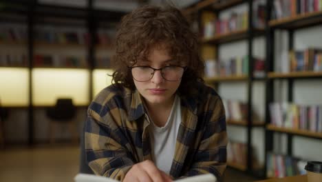 confident girl student with glasses with curly hair reads a book at a table in a university library