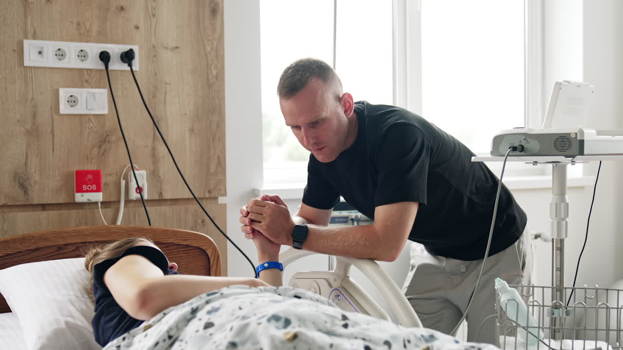 Concerned man holding a hand of his pregnant wife lying in hospital bed. Husband supporting his wife at contractions.
