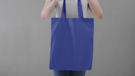 Mujer-Caucásica-Con-Camiseta-Blanca-Sosteniendo-Una-Bolsa-Azul-Sobre-Fondo-Gris,-Espacio-Para-Copiar,-Cámara-Lenta