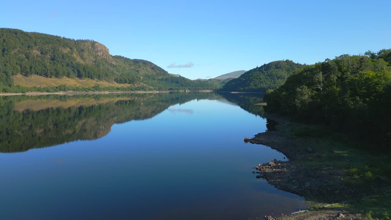 Flying low over calm reservoir with rise revealing woodland and mountains beyond on sunny summer morning at Thirlmere, English Lake District, Cumbria, UK