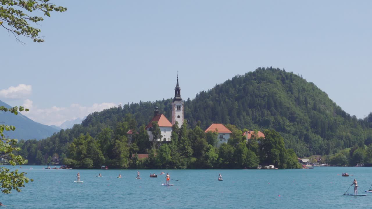 Uitzicht op het eiland Bled in het meer van Bled vanaf de oever met veel stand-up-paddle-boarders op de voorgrond.