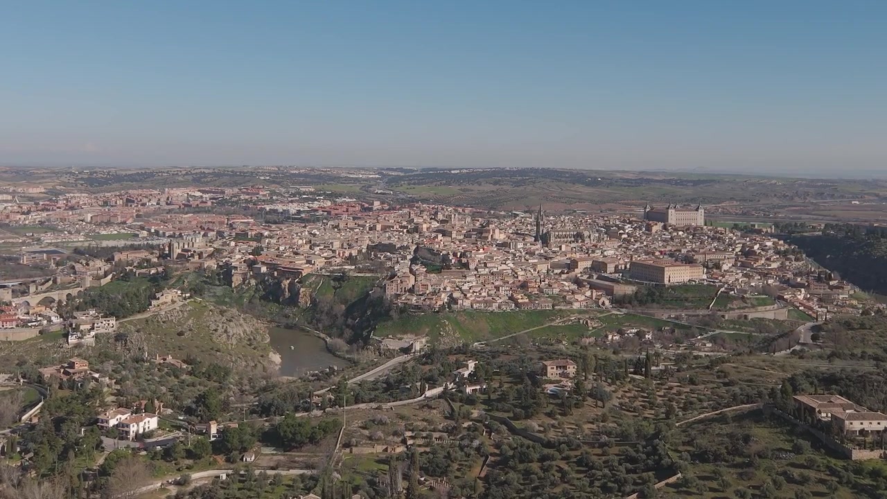 Volo frontale con drone dalla verde zona dei Cigarrales verso la storica città di Toledo. L'Alcázar, la Cattedrale e un tratto del fiume Tago risaltano sotto un cielo azzurro e limpido