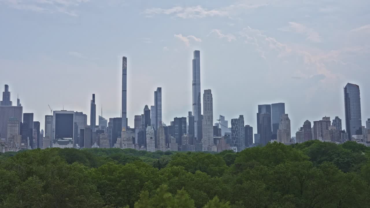 A wide shot of New York City's iconic skyline viewed from Central Park, with towering supertalls rising above the lush green canopy on a hazy summer day.