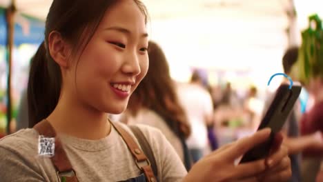 a joyful woman engages with her smartphone as she scans qr codes at a lively outdoor market. the atmosphere buzzes with people enjoying the day.