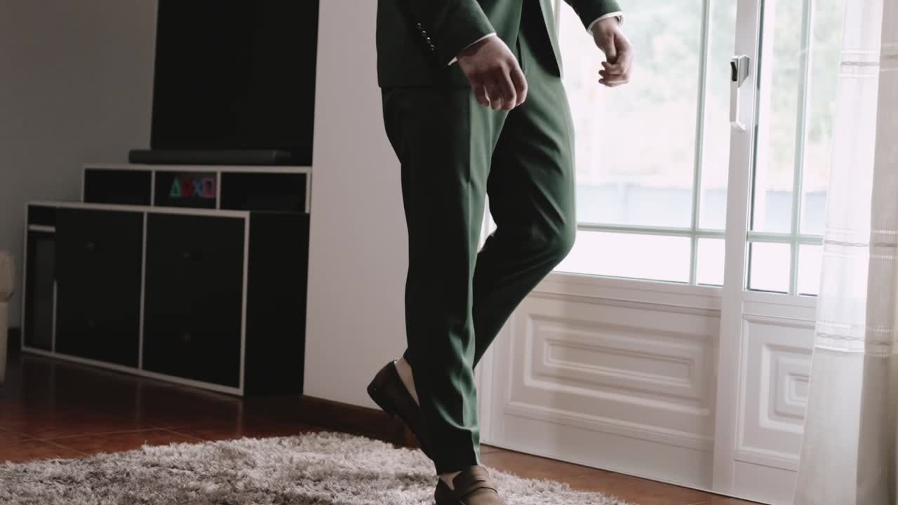 Man in a green suit walking indoors on a plush carpet near a large window, in a modern room