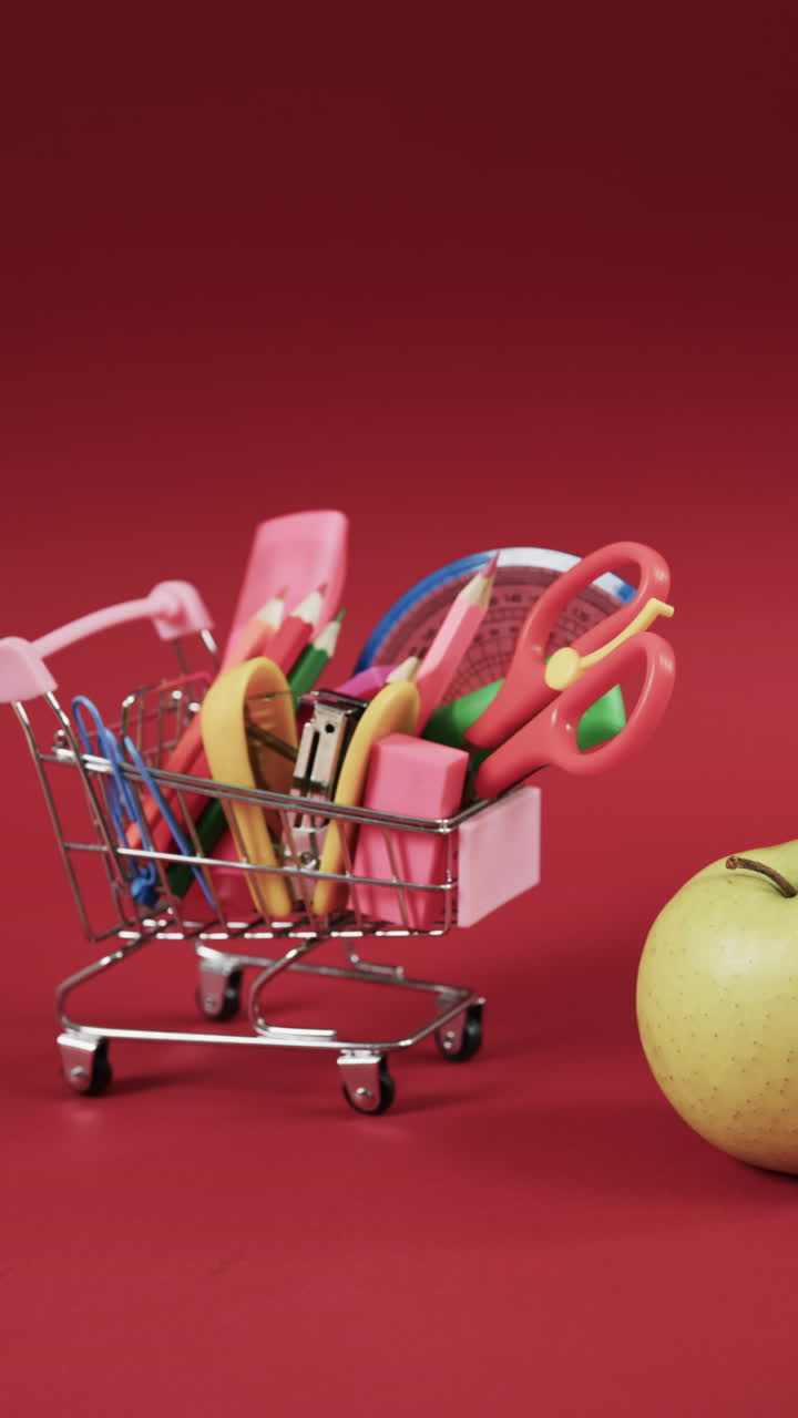 Vertical video of shopping trolley with school materials, apple and copy space on red background