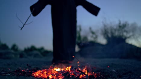 spooky death in black gown near fire. embers of fire on the ground and mysterious figure doing shamanism with a wooden stick. halloween day.