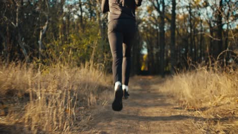 A-male-athlete-in-a-black-sports-uniform-and-black-sneakers-runs-along-the-ground-along-a-low-narrow-path-among-dried-grass-and-tall-trees-in-the-autumn-forest-and-parks