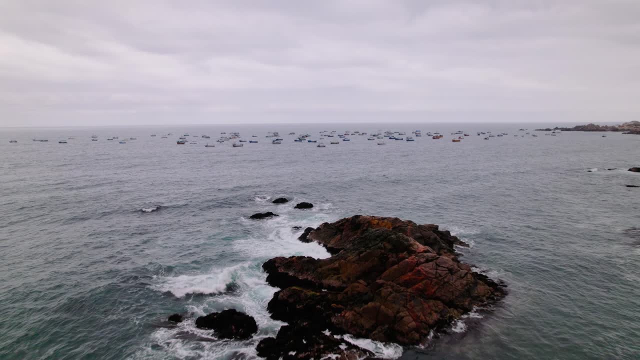 Aerial zoom in of a rocky island surrounded by fishing boats on the Peruvian coast under cloudy skies