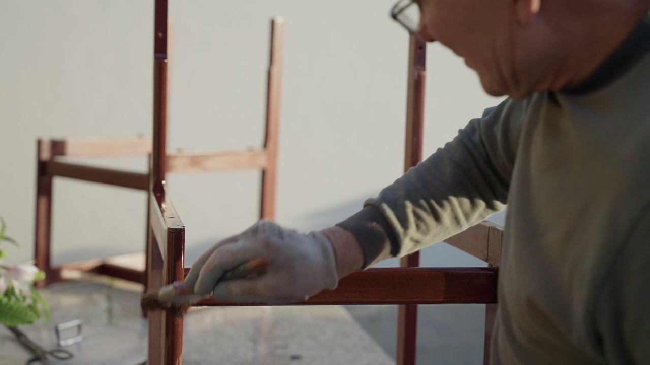 Person in work gloves applying varnish or stain to wooden chair frame during restoration