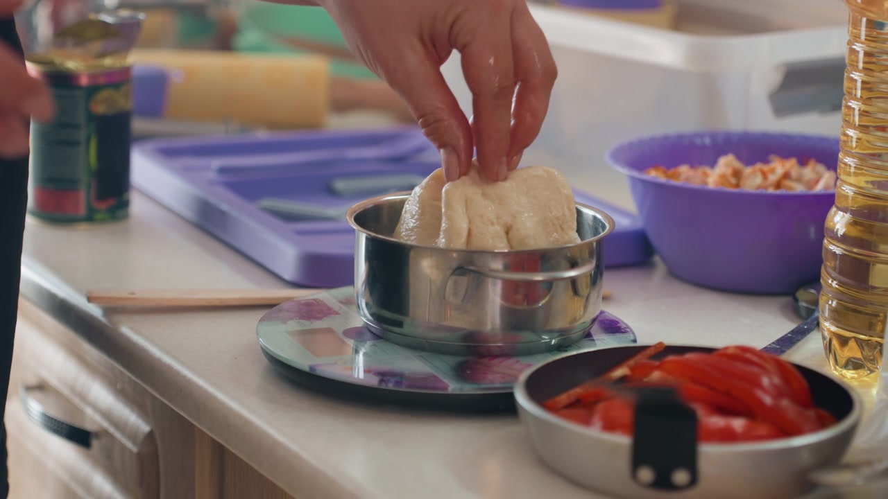 Close-up kitchen workspace with metal sifter holding pressed ingredient on digital scale beside bowl of sliced red peppers and clear containers, showing careful prep, organized tools, and subtle