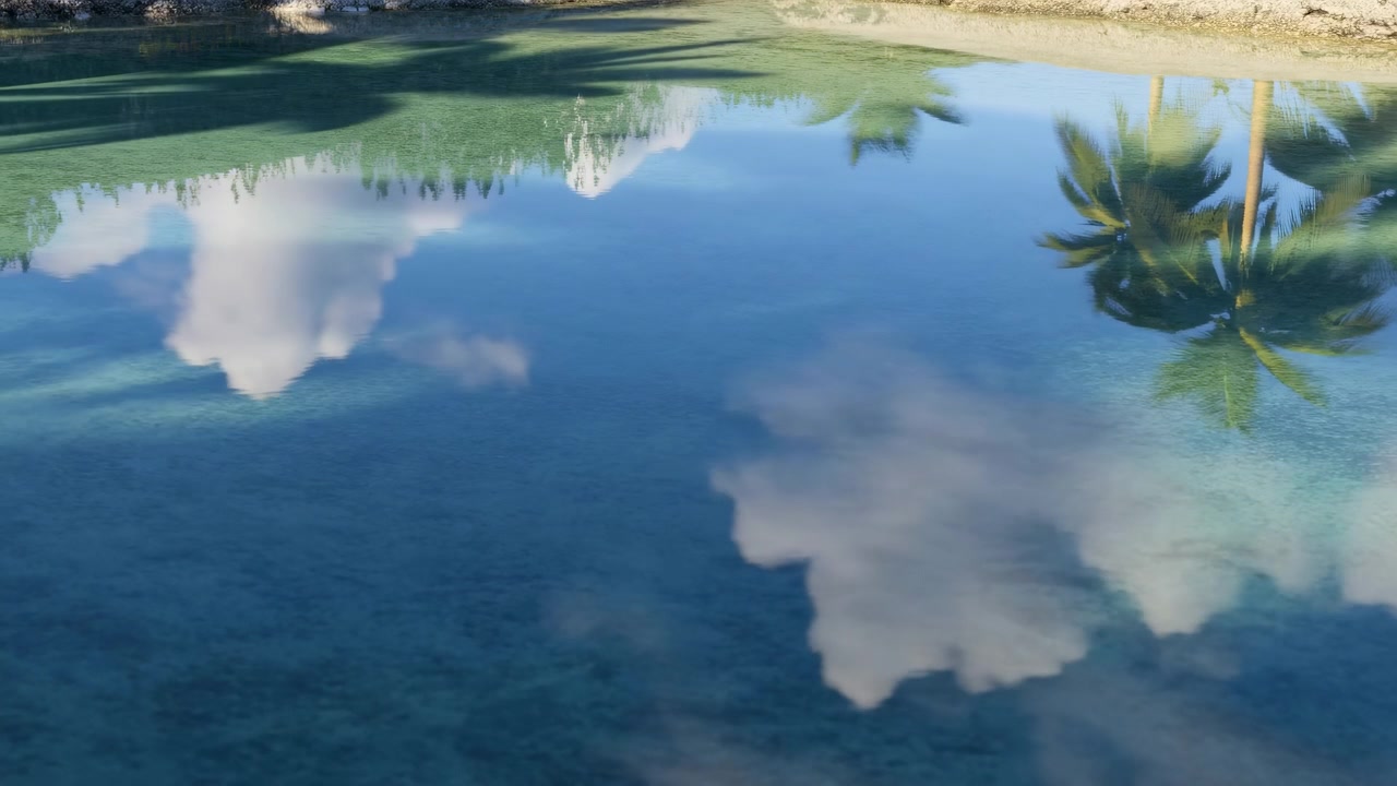 Rustige tropische lagune met spiegelende wolken, palmbomen en zandige kustlijn, waar subtiele, verschuivende reflecties over glanzend blauwgroen water bewegen onder helder daglicht en een vredig kustlandschap.