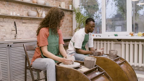 employees wearing green apron modeling ceramic pieces on potter wheel in a workshop while talking to each other 1