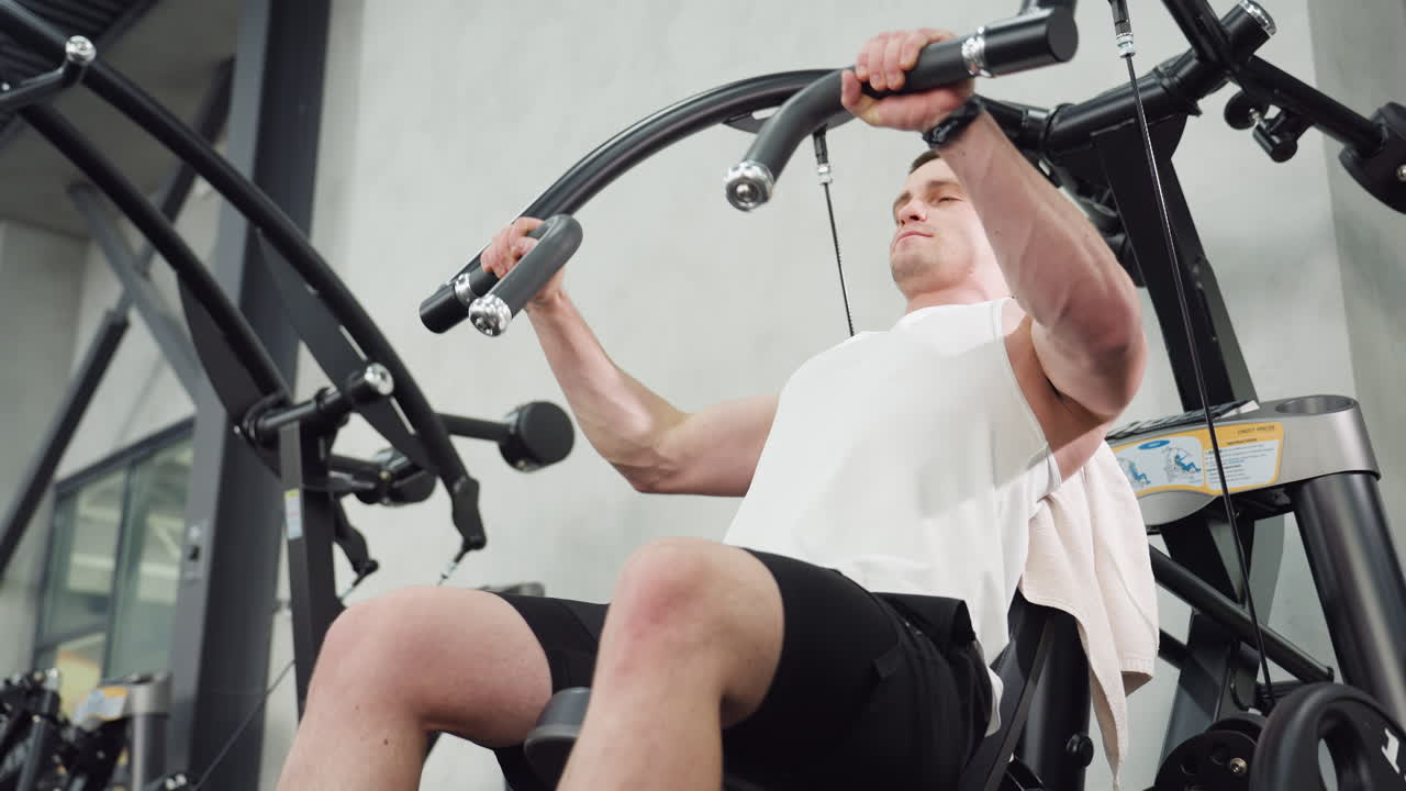 Keep fit buff working out on chest press machine hands gripping curved handles eyes closed face tightened muscles straining under modern gym lighting on sleek black steel equipment intensity