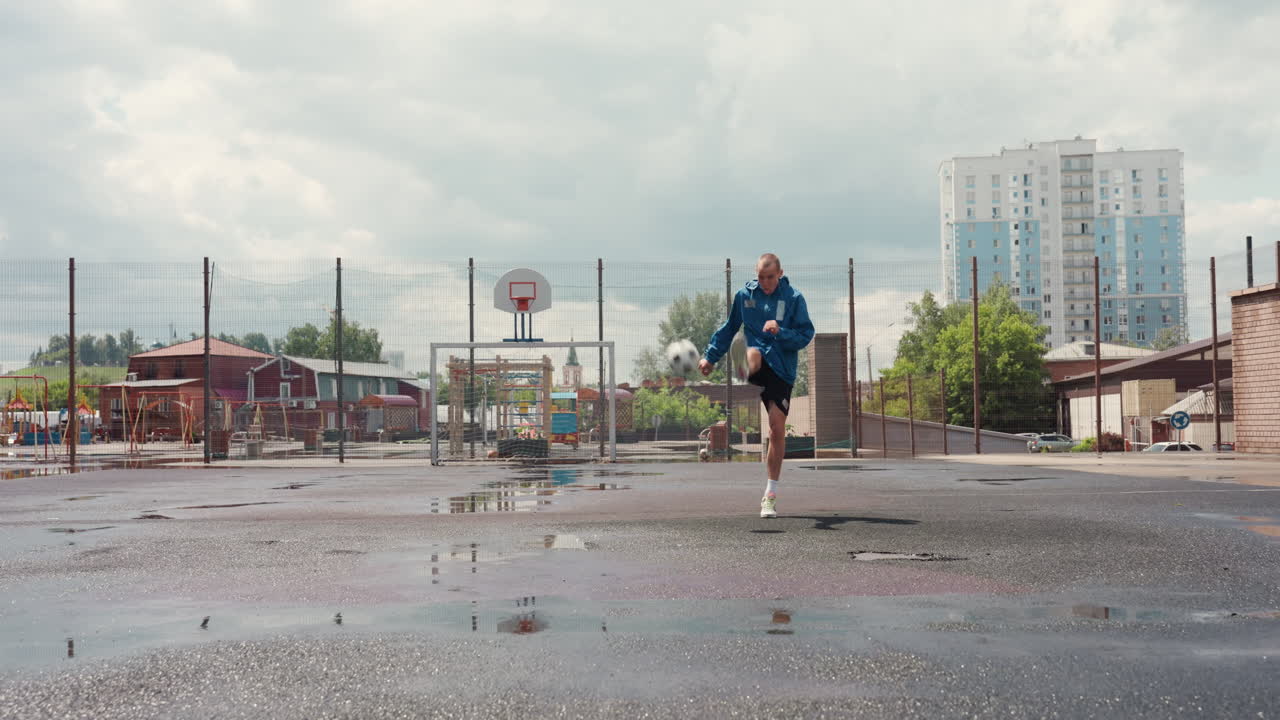 giocatore che palleggia su un campo bagnato sotto un cielo plumbeo, atleta solitario in giacca blu che si allena a calcio su un pavimento zuppo di pioggia, pozzanghere che riflettono il profilo distante della città e i grattacieli, recinzione metallica