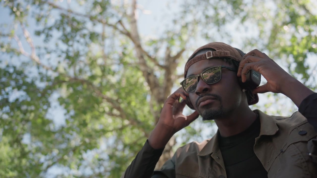 Close up of man wearing brown cap and jacket adjusting over ear headphones in outdoor setting with green trees and blue sky. Man inspects headphone details focusing on device interaction