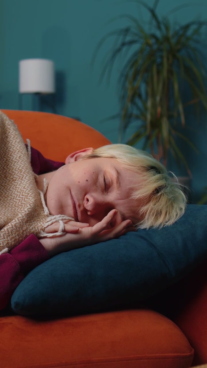 Tired blonde young woman lying down in bed taking a rest at home napping falling asleep on sofa