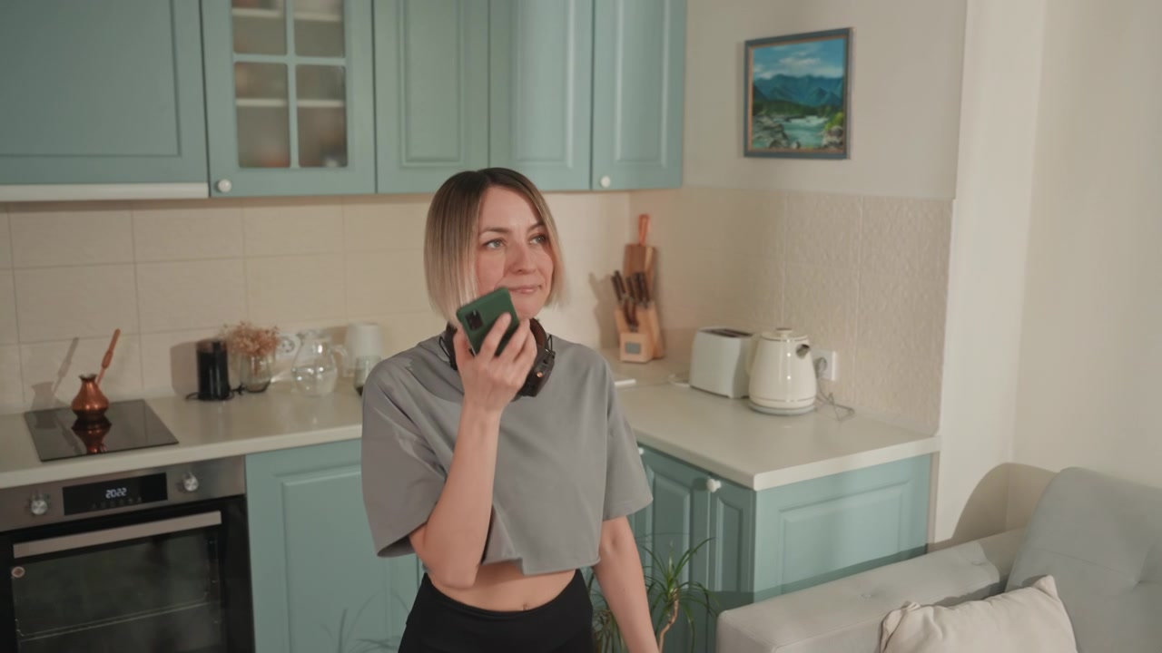 Donna bianca che sfoglia una playlist sul telefono, cuffie appoggiate al collo, scorrendo un'app di streaming, luce solare che inonda una cucina dai toni pastello, atmosfera mattutina rilassata, profumo di caffè, sorriso sottile, scoperta di contenuti.
