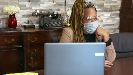 black woman in glasses wearing mask sitting in office in front of computer gazing out of the window daydreaming