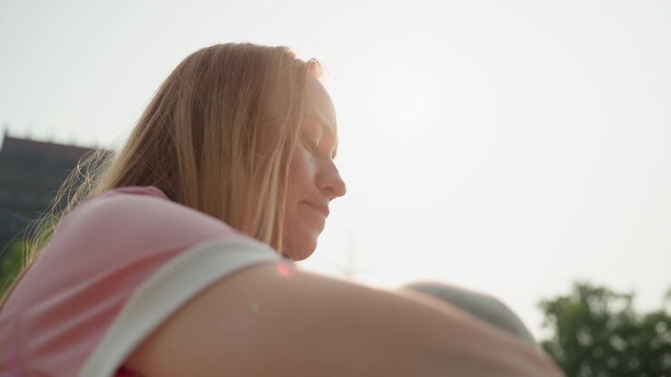 Woman with long blonde hair wearing pink shirt is seen from side her profile visible as woman looks downward. background features urban setting with blurred buildings and greenery under soft