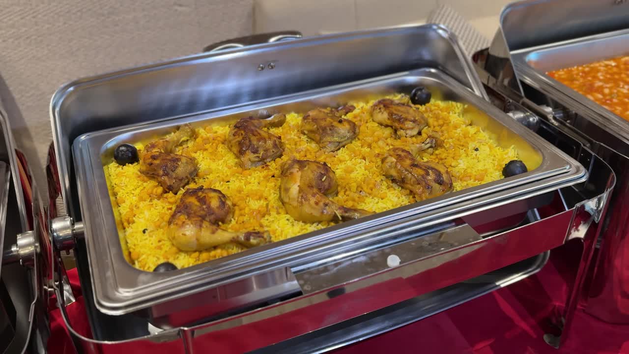 Buffet da catering al chiuso con scaldavivande in acciaio inossidabile che serve cosce di pollo grigliate su riso giallo speziato con olive nere, esposizione di cibo caldo su un tavolo da banchetto sotto un'illuminazione calda