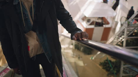 close-up of man's hand resting on escalator railing in modern shopping mall, casual outfit with hoodie, denim jacket, and small bag over shoulder, soft lighting in bright interior