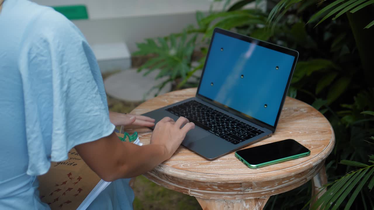 un joven europeo en un vestido azul remotamente en línea trabajando en una computadora portátil con pantalla azul en el patio trasero con plantas verdes