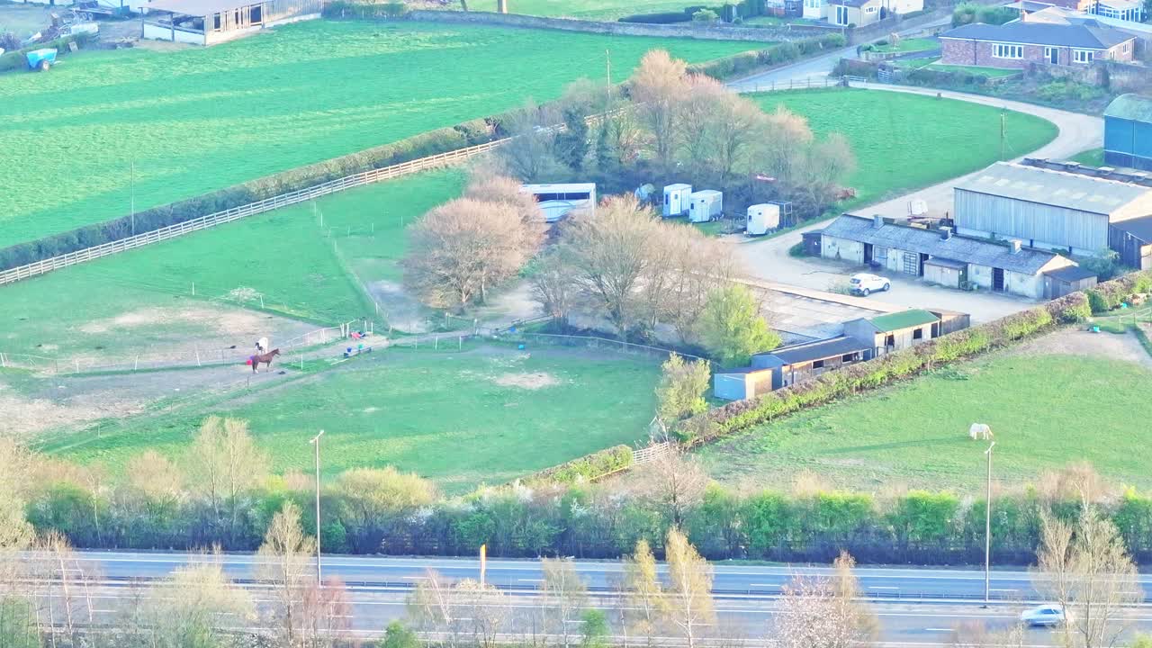 Aerial drone shot over rural horse paddock, grazing horses, stables, and nearby fields with farm buildings near Lundhill, Wombwell, South Yorkshire, England