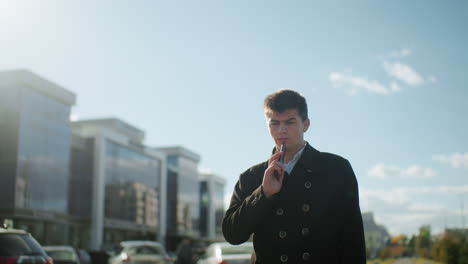 man wearing black coat on phone call outdoors under clear blue sky, walking near parked cars and modern glass buildings, appearing displeased and contemplative during conversation