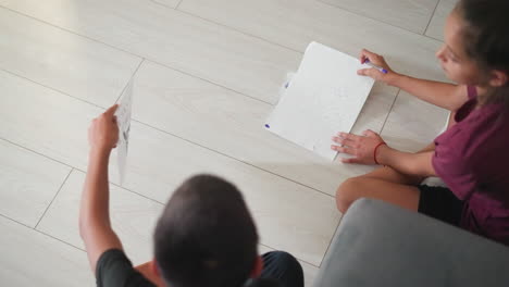 siblings seated on wooden floor busy writing and sketching side by side boy shows his work to sister while she shares hers too capturing creative exchange learning interaction