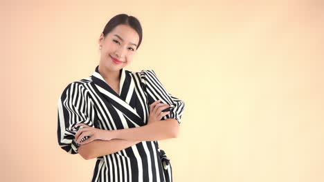 Pretty-stylish-asian-business-woman-smiling-to-camera-and-crossing-her-hands,-full-frame-slow-motion,-isolated-on-beige-background