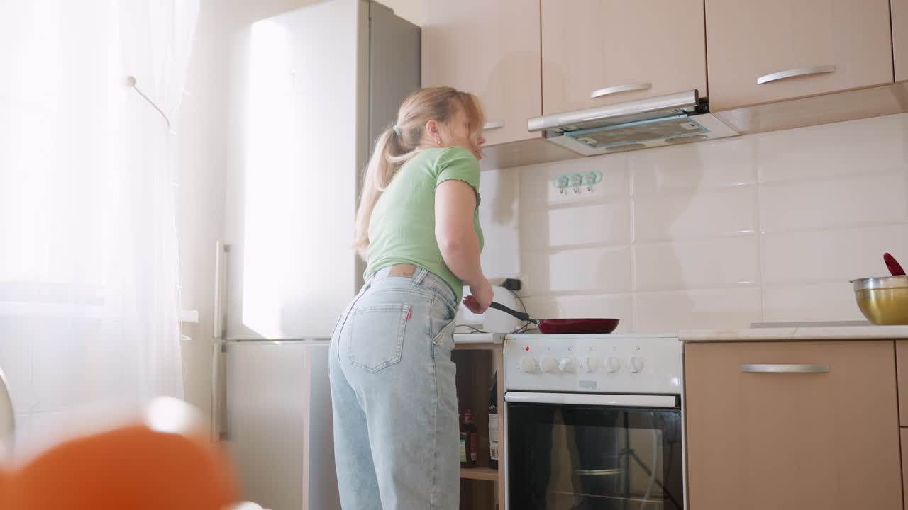 Woman in green shirt and jeans bends near stove collecting cooking oil bottle from lower cabinet and places it on counter during food preparation in bright modern kitchen
