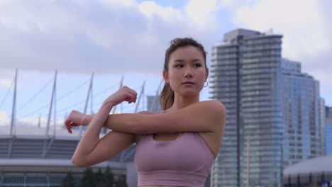 mujer estirando tríceps con el brazo cruzado, edificios de la ciudad en segundo plano.