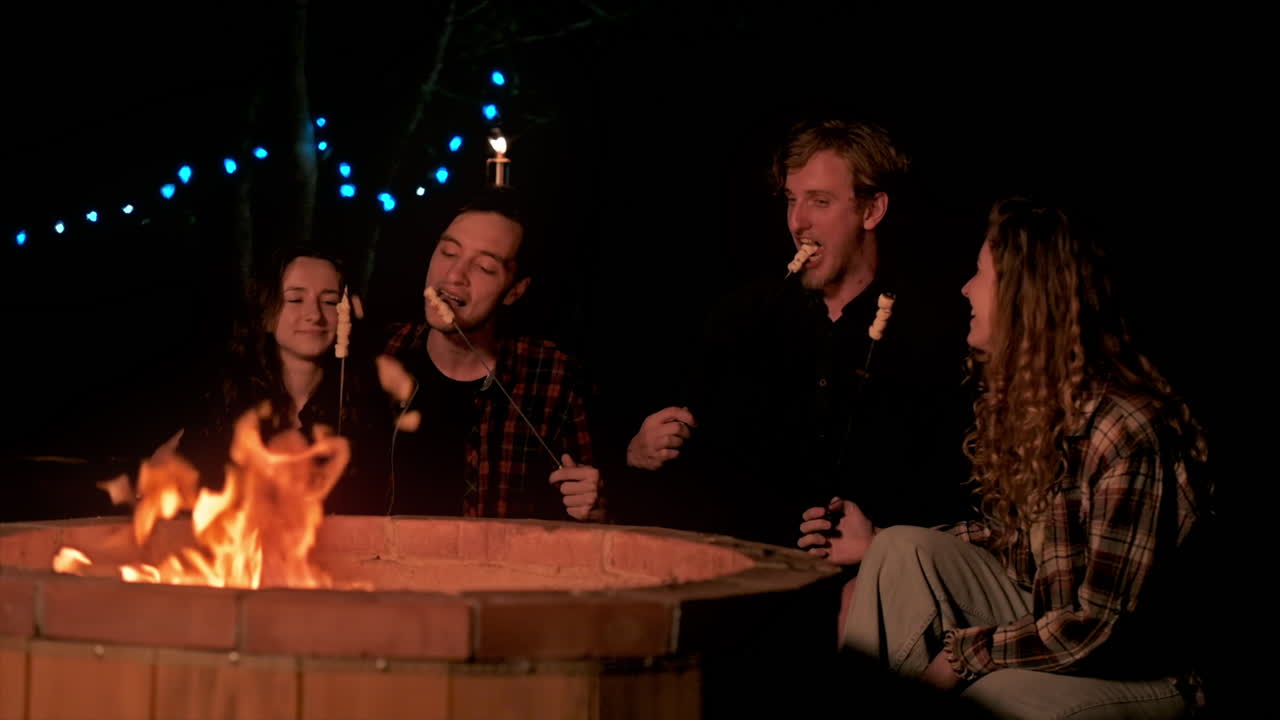 A group of happy young friends near a campfire at glamping, night. Two men and women. Frying and eating marshmallow. Slow motion