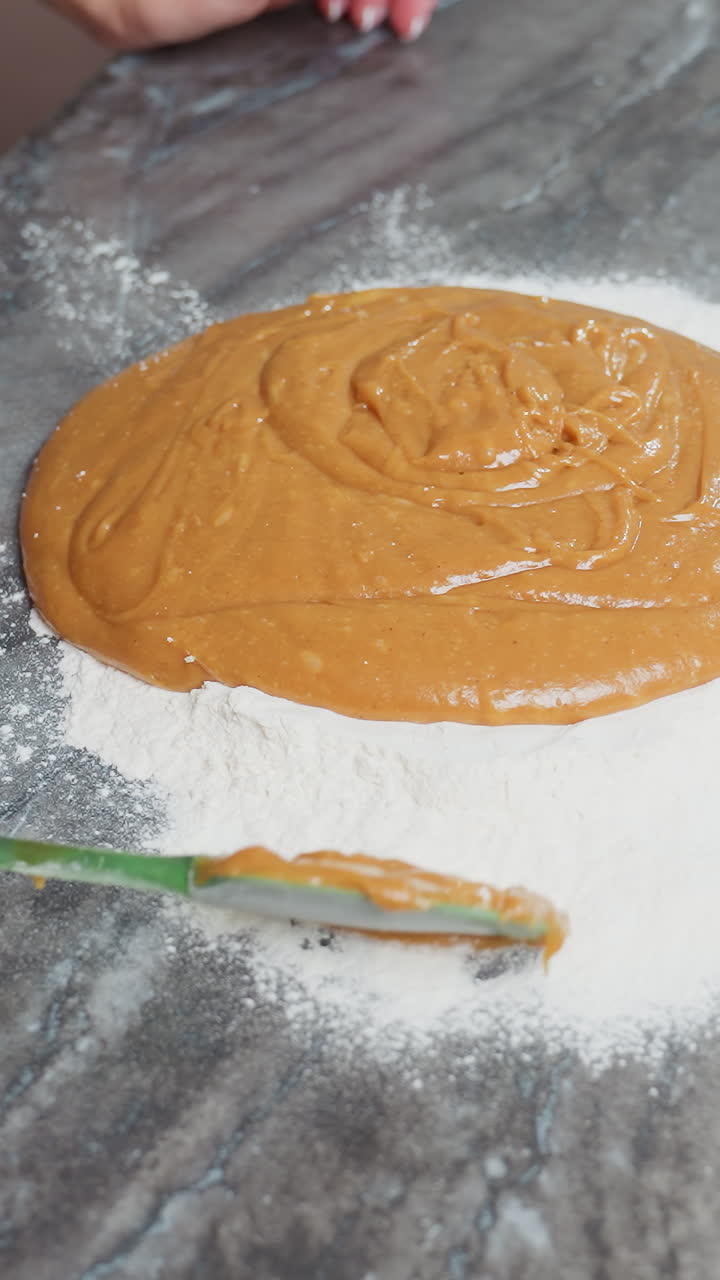 Aerial view of woman using green spatula to cover thick brown dough with flour on a clean countertop, baking preparation in a cozy kitchen, focusing on hands