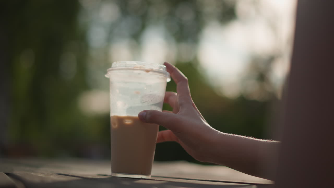 Giovane donna allunga la mano verso un caffè freddo, studentessa si ferma su un patio di legno durante l'ora d'oro, ripresa al rallentatore, bicchiere di plastica con condensa, calda retroilluminazione e bokeh delicato, rilassata solitudine estiva