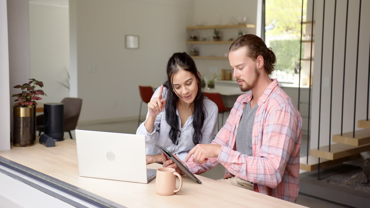 Young happy couple smiling using laptop and tablet together in kitchen at home