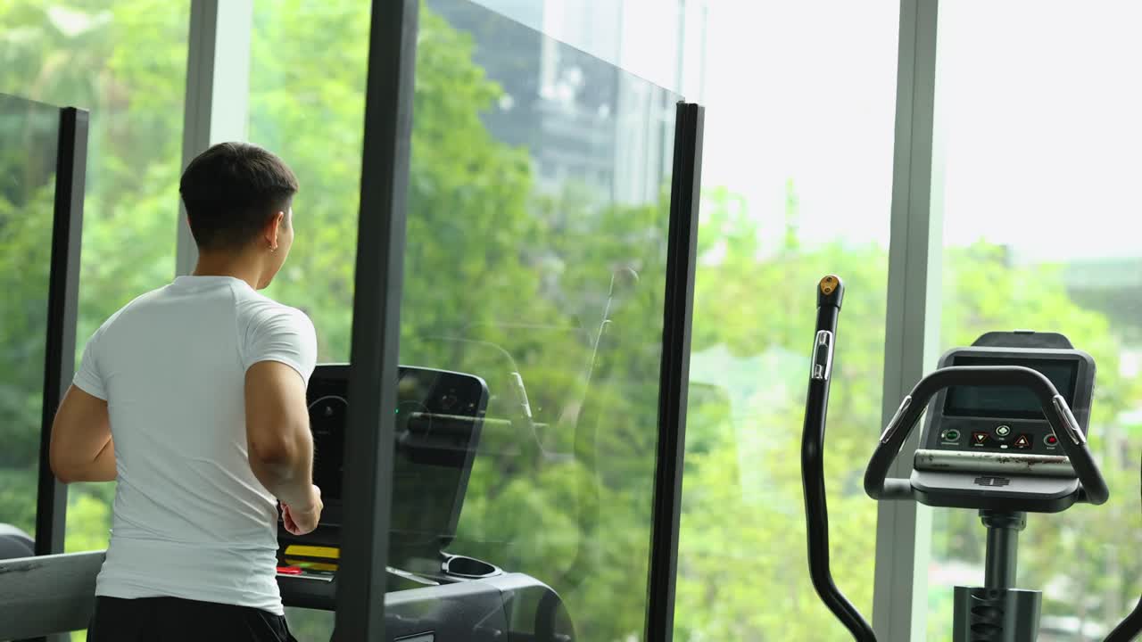 Man running on treadmill in bright modern gym with large windows overlooking greenery in Bangkok