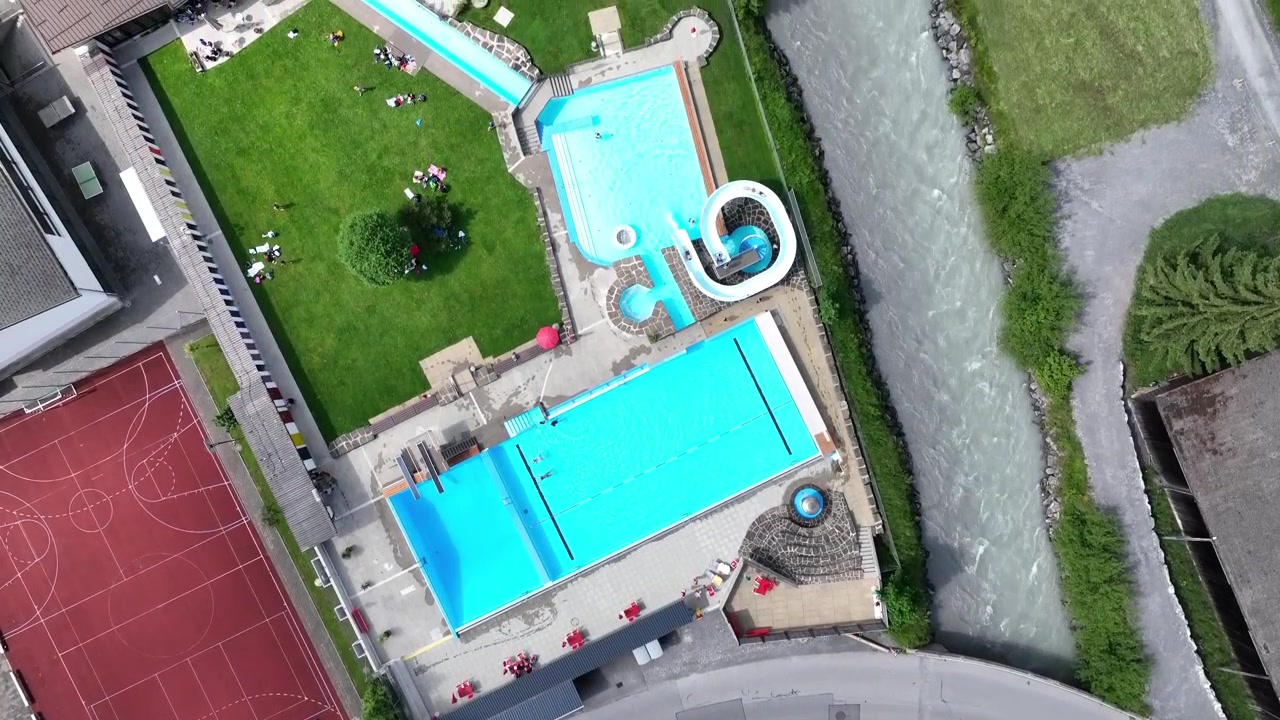 Luftoptagelser, der viser en livlig schweizisk udendørs swimmingpool med en vandrutsjebane, en rød sportsbane, frodige grønne omgivelser og en nærliggende strømmende bæk i dagslys, Schweiz