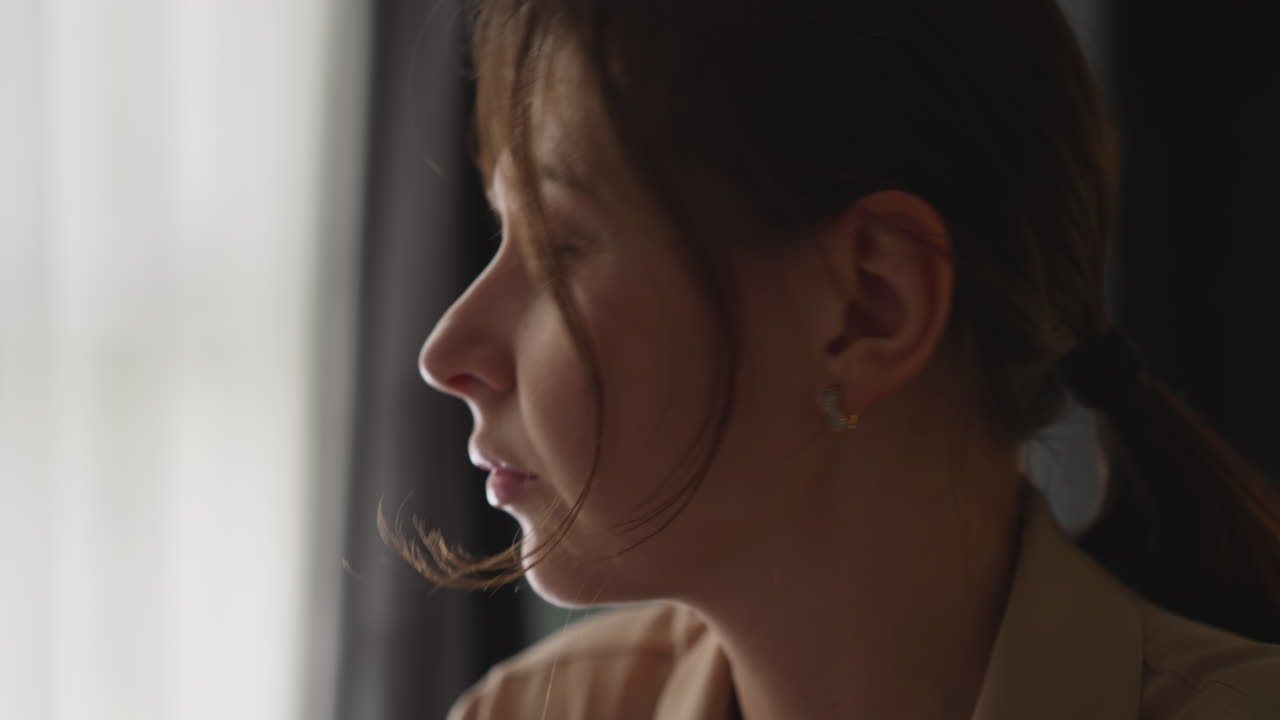 사려 깊은 여자가 창문에서 성찰하고 카메라를 쳐다본다.