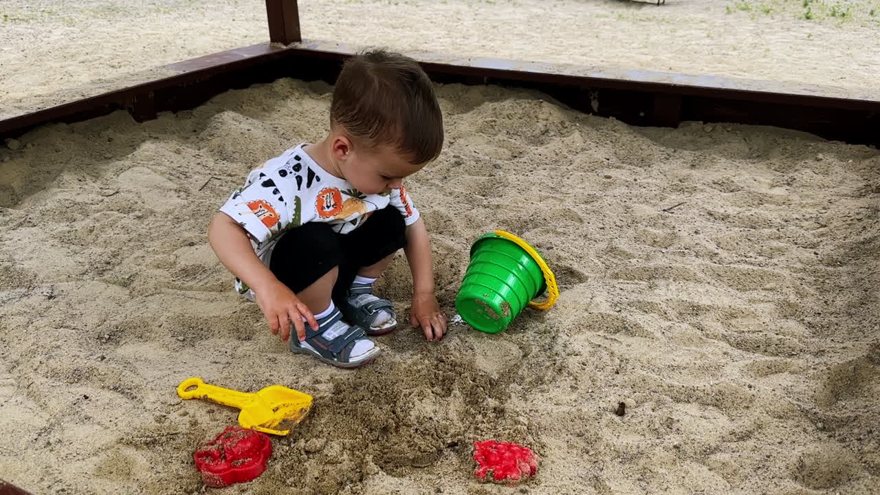 Little Caucasian kid sitting in the sandbox poured water from his bucket. Baby boy touches the wet sand looking pensively away.